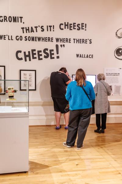 A gallery wall with large black text reading “Gromit, that’s it! Cheese! We’ll go somewhere where there’s cheese!” attributed to Wallace. Below the text are framed sketches and a display case with a small robot figure. Three visitors stand reading information panels.