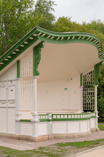 Victorian-style outdoor bandstand — a small white and green wooden performance pavilion with a sweeping curved roof and decorative brackets, set on a paved area beside a tree-lined park.