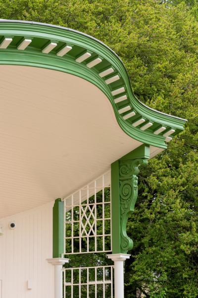 Ornate green-painted wooden cornice and carved bracket supporting a curved porch eave with cream beadboard ceiling and white dentil blocks, set against leafy tree branches.