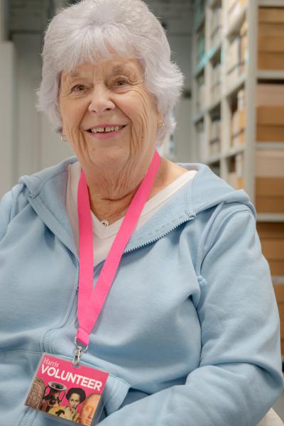 An elderly lady in a blue jumper and a pink lanyard sat looking at the camera in a back room setting