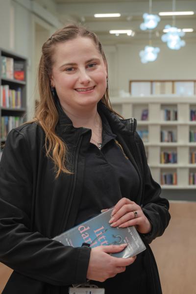 A young woman with long hair in a library setting, holding a book looking at the camera and smiling.