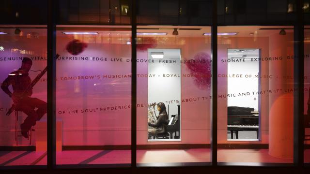 Street-level view of a modern conservatoire. A person can be seen playing an instrument through the window.