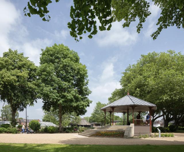 a woman pushes a pram past a bandstand in a park