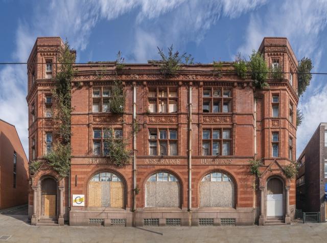 a brick building with three large ground floor arched windows and buddleia growing from the facade