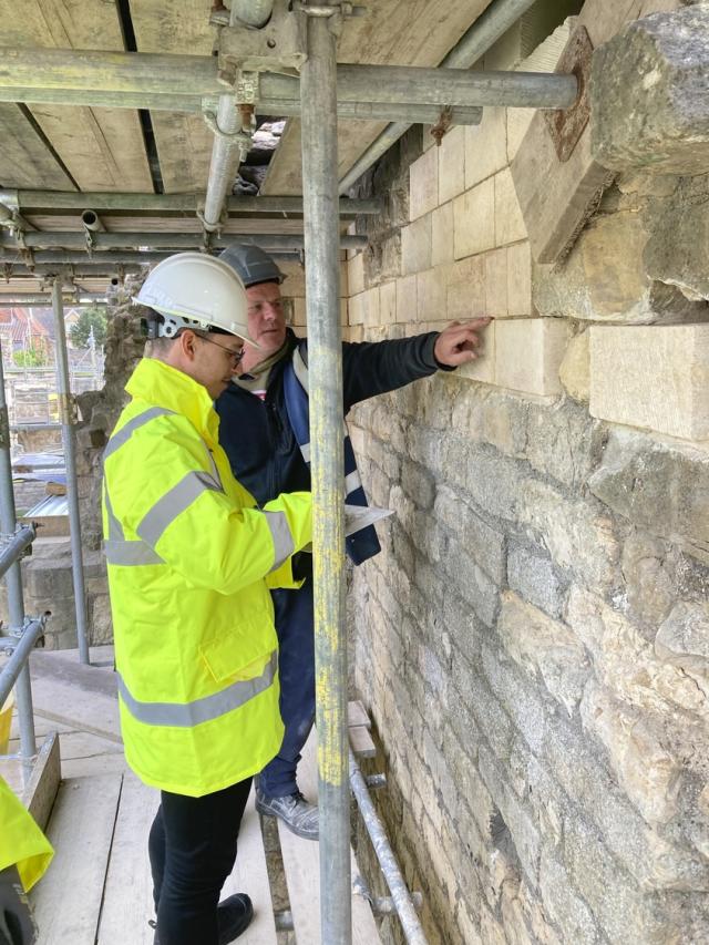 a stone mason in a high visibility jacket is working on a medieval wall - behind him is a man pointing to the pointing in the wall.  They are standing on scaffolding. 