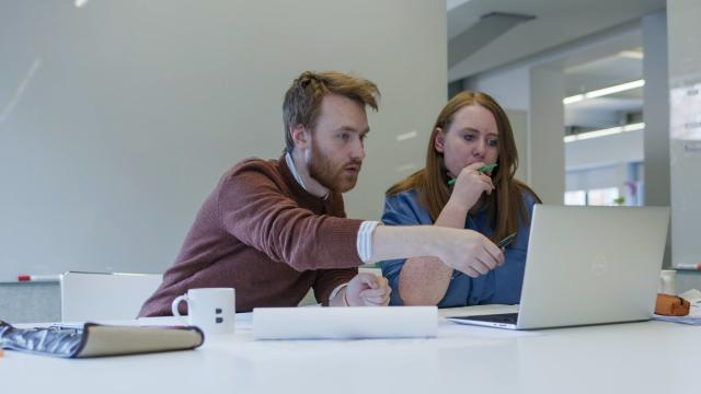 a man in a brown jumper and lady in a blue shirt discuss designs on a laptop