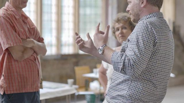 a man in a checked shirt is describing something to a man in a pink shirt and lady in a white blouse at a consultation event