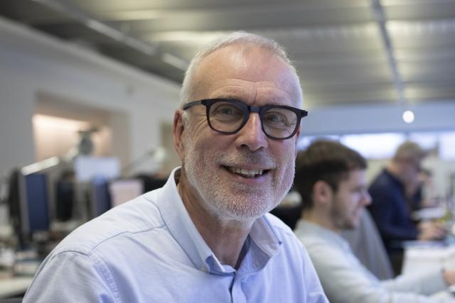 Photograph of a man smiling at a camera. He is in an office setting. He is wearing black rimmed glasses and has silver hair.