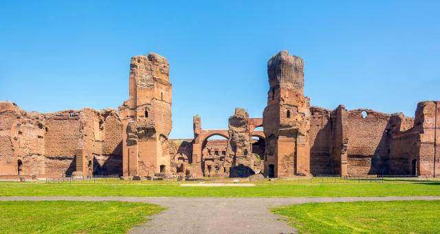 External landscape photograph. In the foreground there is neat green grass and a pathway leading to pink/red coloured stone building in ruins. There appear to be two ruined towers and walls.  