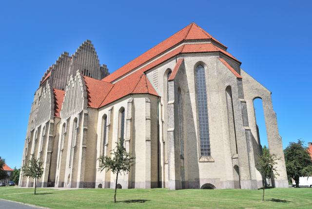 External photograph of a modern grey church with a red tile roof. The front facade of the church has three triangulated pinnacles reaching towards the sky. There are two flying buttresses at the end closer to the viewer. The church sits in a green lawn with trees. 