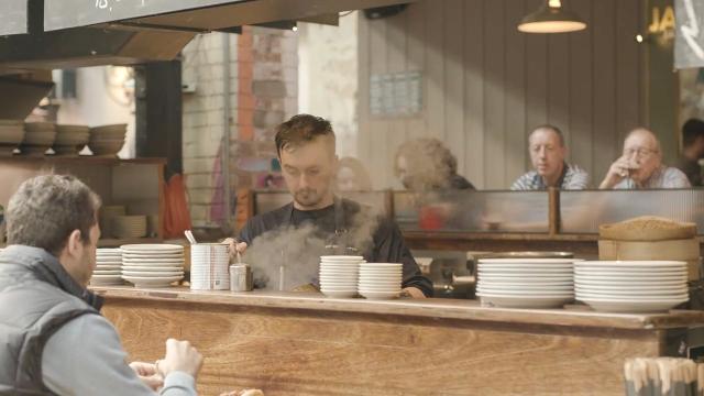 an open kitchen at the mackie mayor, a wooden bar with bowls stacked on it is in front of a chef with steam rising in front of his face
