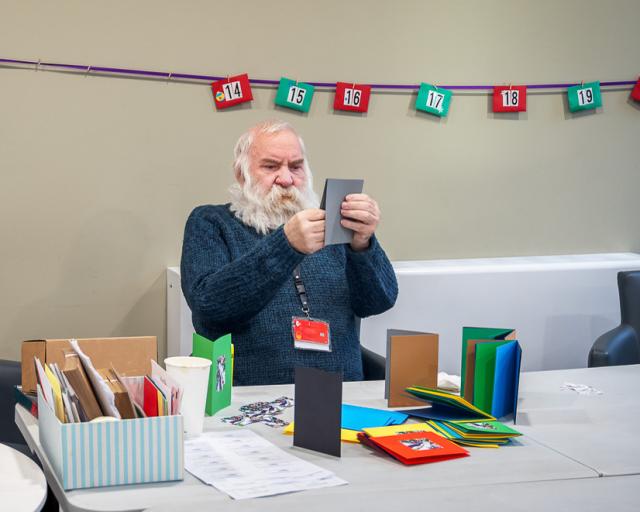 a man with long white beard and in a blue jumper is making cards, there is a red and green bunting calendar on the wall behind him