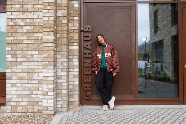 a lady in a brown bomber jacket stands in front of the greenhaus sign on the front door of the new apartments