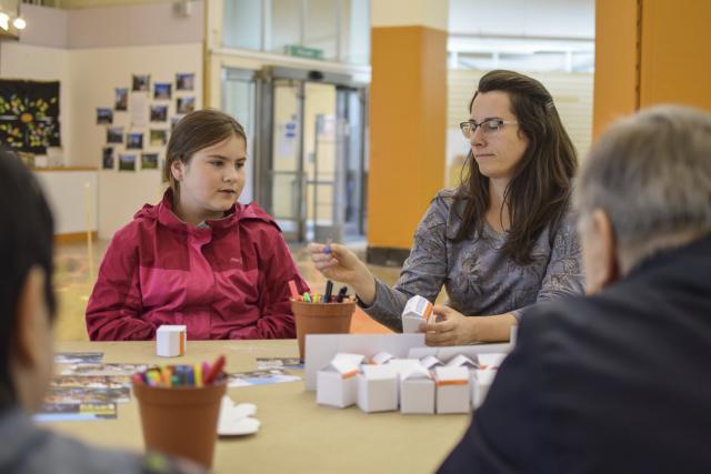 A woman and a girl sit at a table engaged in an art activity with another person. The table is covered with art supplies and small white boxes.