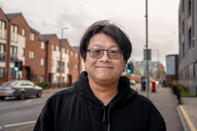 a man wearing a black hooded top stands on a city centre street. he is smiling at the camera.