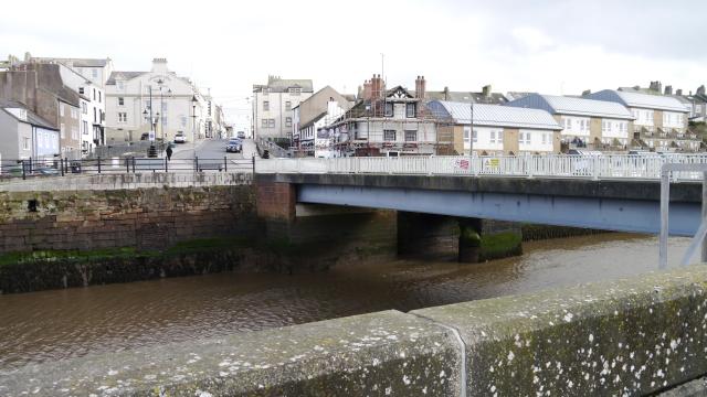 a blue bridge spans the river in maryport with white and brick buildings facing from the other side