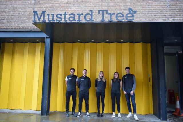 Group of people standing in front of a yellow metal wall with Mustard Tree sign above it