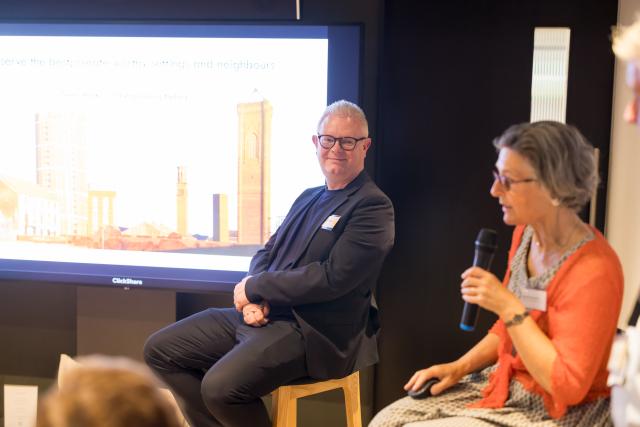 A woman holding a microphone in front of a presentation screen. A man sits next to her.