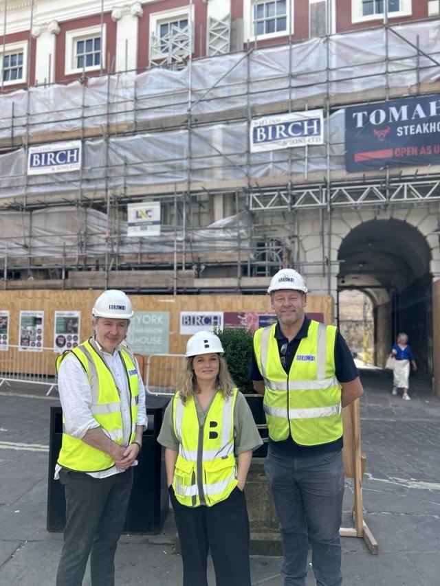 Three people in hard hats and high vis vests standing outside a building covered in scaffolding.
