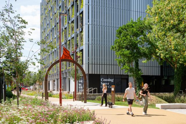 Modern building with integrated greenery, a flower-lined path, two people walking—one with a dog—and an archway sculpture; 'Coming Soon' sign visible.