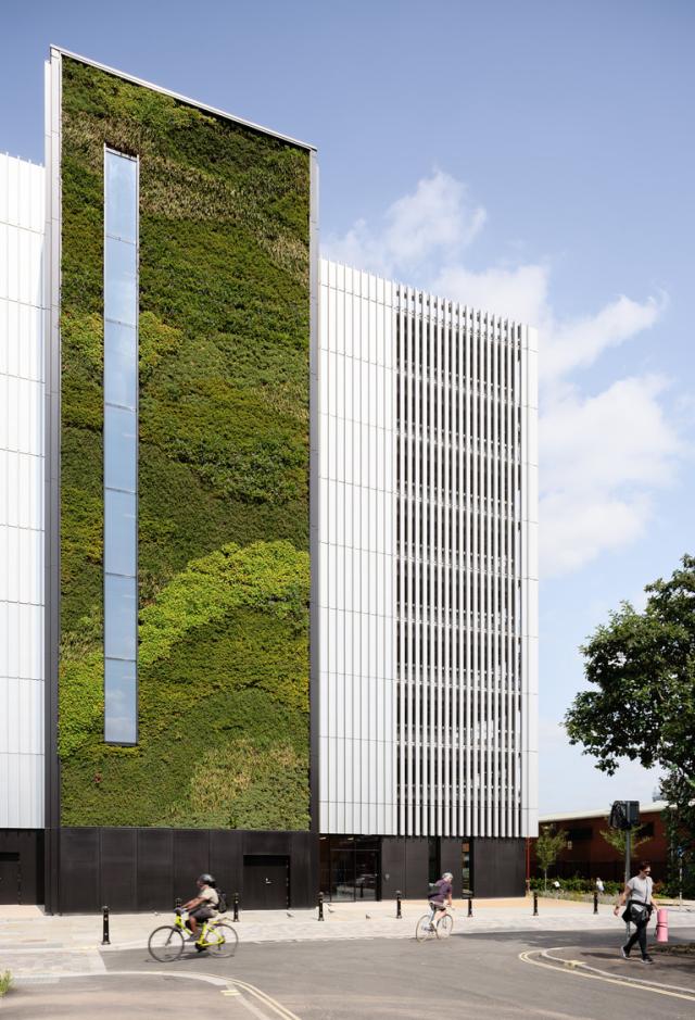 Modern building with a vertical garden, glass windows, and people walking and cycling in an urban setting with a crane in the background.