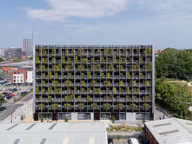 Multi-storey building with vertical greenery on its facade, surrounded by buildings, trees, and parked cars.
