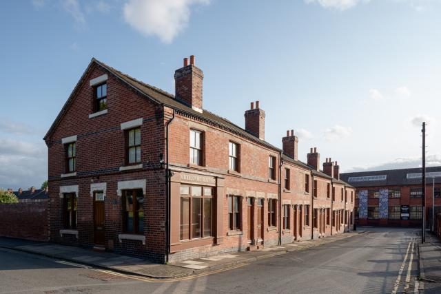 Street-level view of a corner red-brick building labeled “Harper Street” on the window, with adjoining terraced houses extending into the distance. The road is empty, and the industrial building with patterned panels is visible in the background.