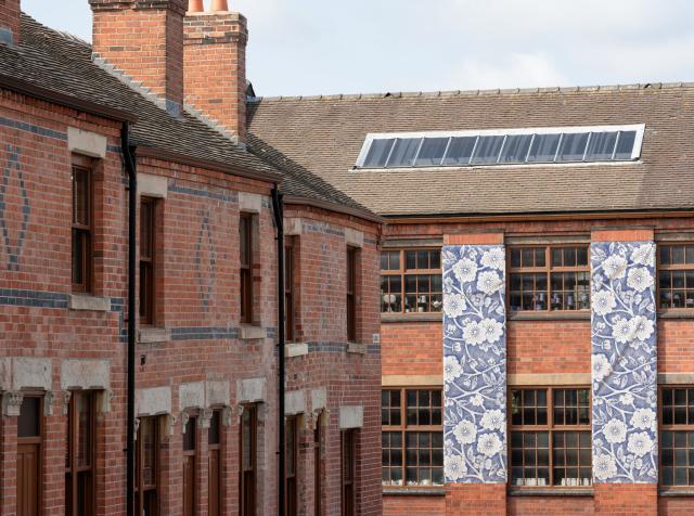 Close-up of two adjoining red-brick buildings: terraced houses on the left and a larger industrial building on the right. The industrial building features large multi-pane windows and two vertical panels with blue-and-white floral designs. A skylight is visible on the roof.