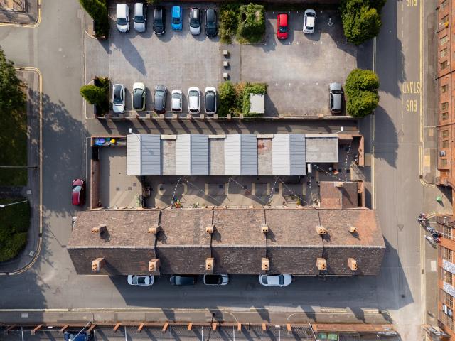 Top-down aerial view of a rectangular block of terraced houses with pitched roofs and chimneys. A courtyard with modern metal-roofed structures runs along the back, and cars are parked on surrounding streets and in a nearby lot.
