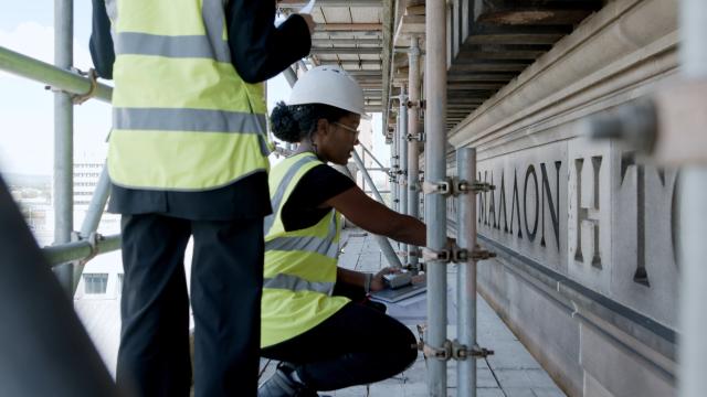 a lady is crouched down looking closely at the inscription on a building from a scaffold