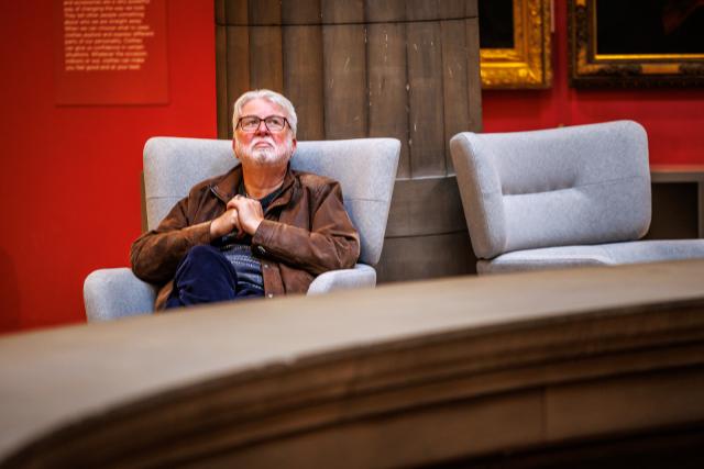 A person sits in a light gray upholstered chair near a large column in a red-walled gallery. Another matching chair is positioned nearby.