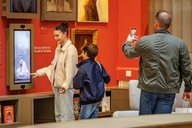 Two visitors stand in front of a vertical touchscreen showing a grayscale portrait, while another person takes a photo. The wall behind is red and decorated with framed paintings and bookshelves.