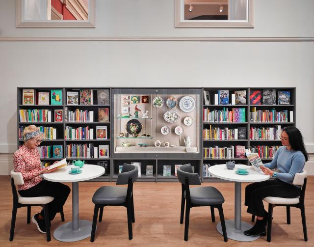 two women sit in front of Tall shelves filled with books in a bright room with classical columns, and a central display case featuring sculptures and artifacts.