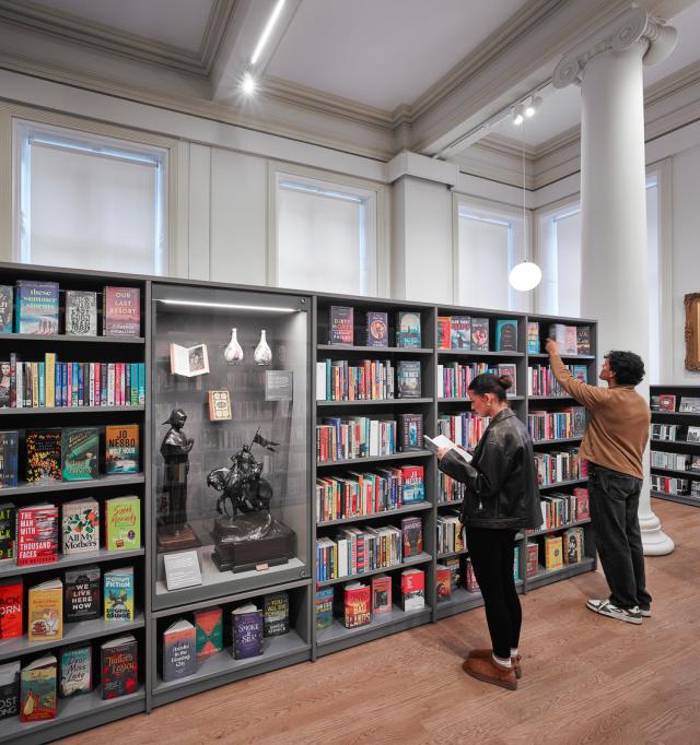 Tall shelves filled with books in a bright room with classical columns, and a central display case featuring sculptures and artifacts.