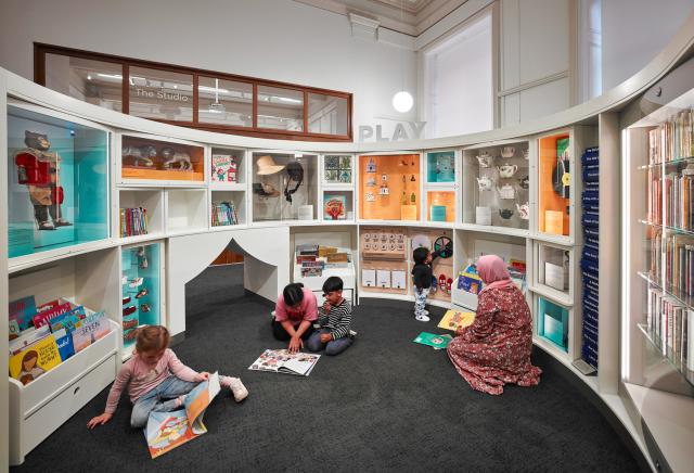 A curved shelving unit filled with colorful books and interactive displays, with children sitting on the floor reading in a bright, modern space.